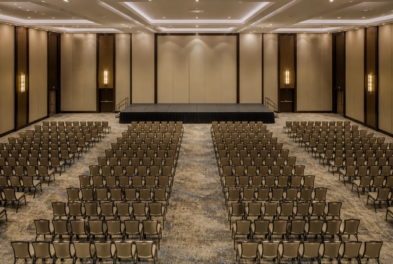 A hall set in theater rows of matched event chairs facing an empty low stage with house lights up.