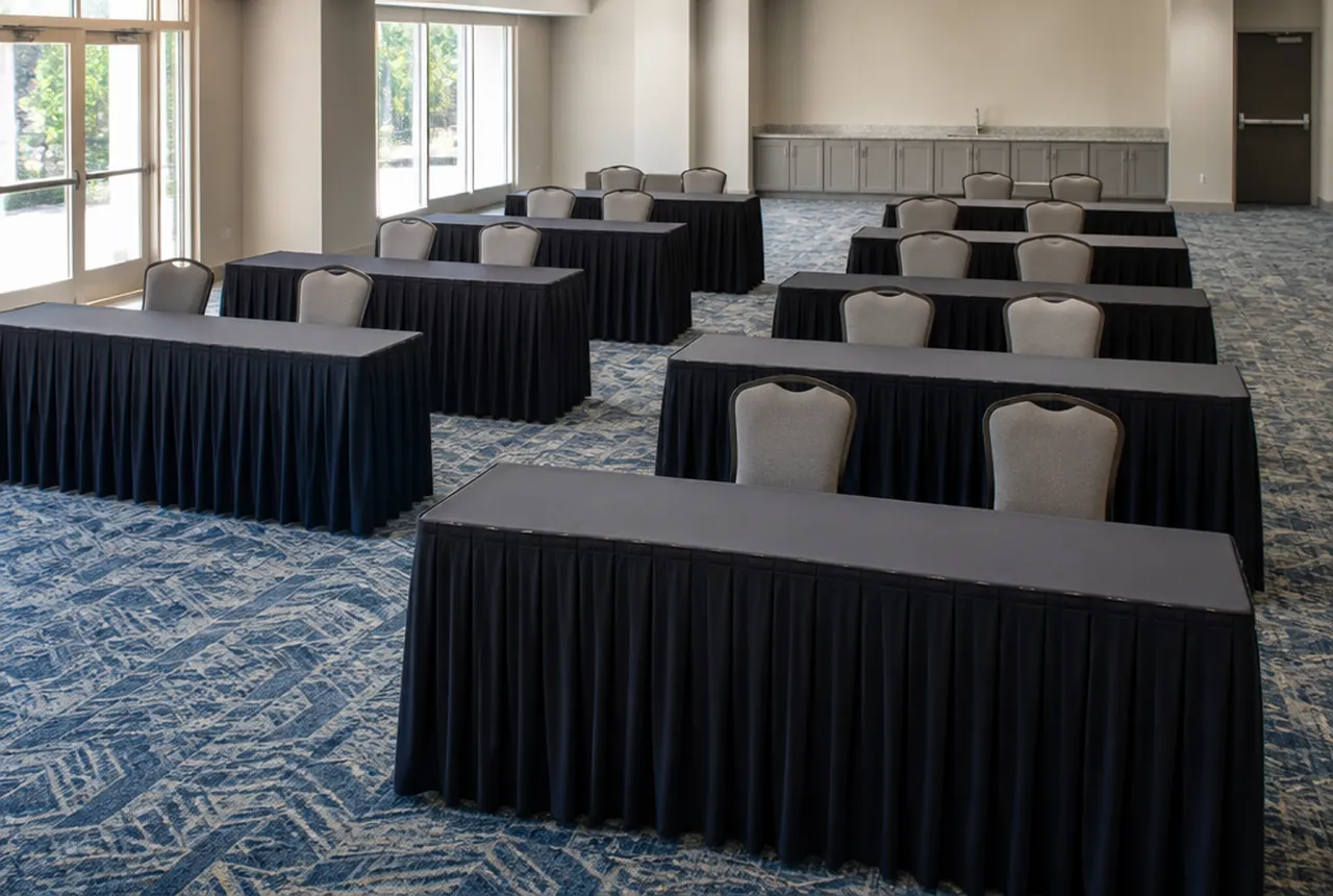 A room set with skirted 6-foot tables in forward-facing rows, two chairs per table and cool daylight.